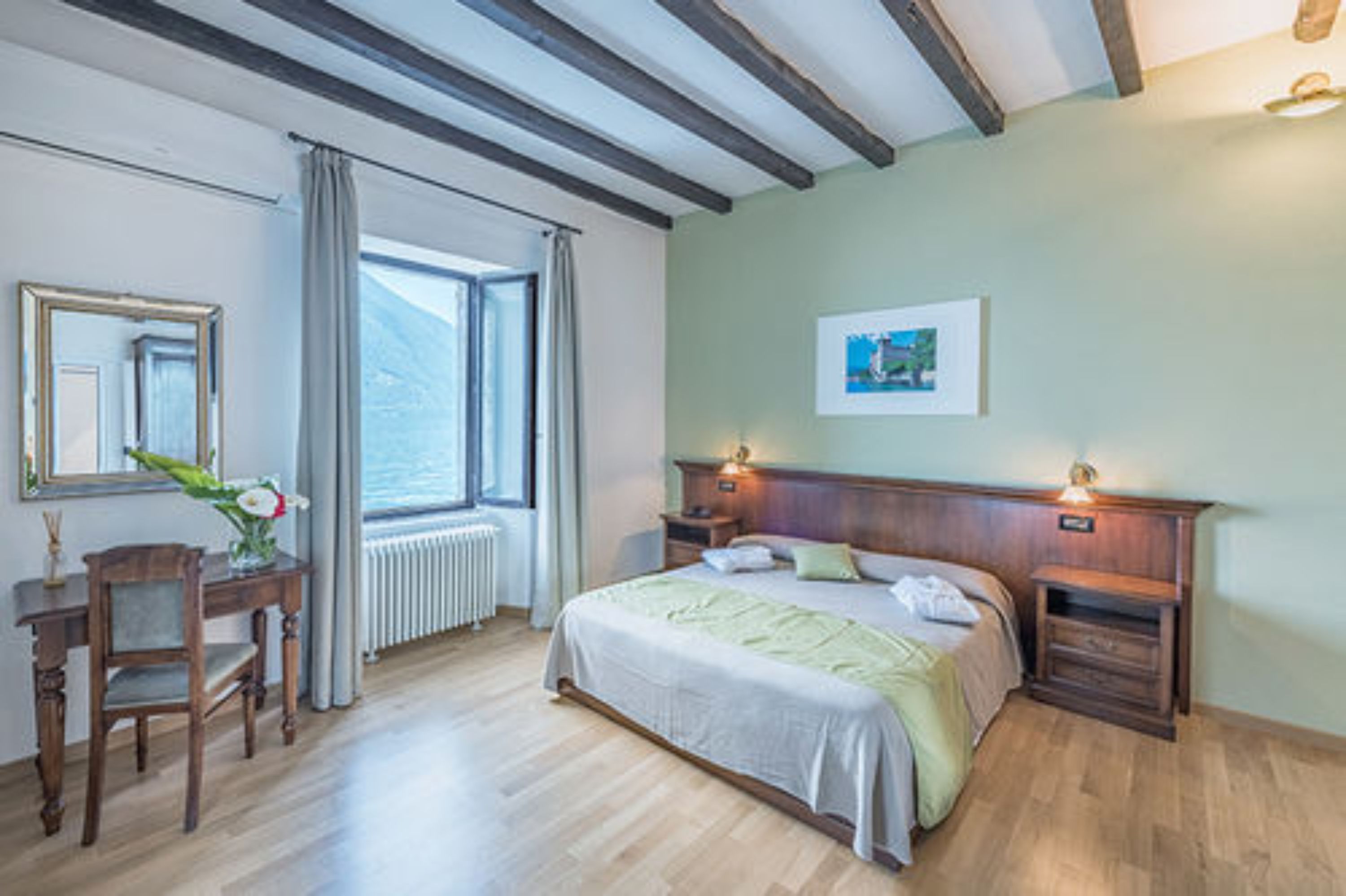 Et soveværelse med en træseng, et natbord, et sengebord, en stol, et skrivebord, en radiator, et vindue med gardiner og et billede i ramme på væggen.