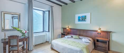 A bedroom with a wooden bed, a nightstand, a bedside table, a chair, a desk, a radiator, a window with curtains, and a framed picture on the wall.