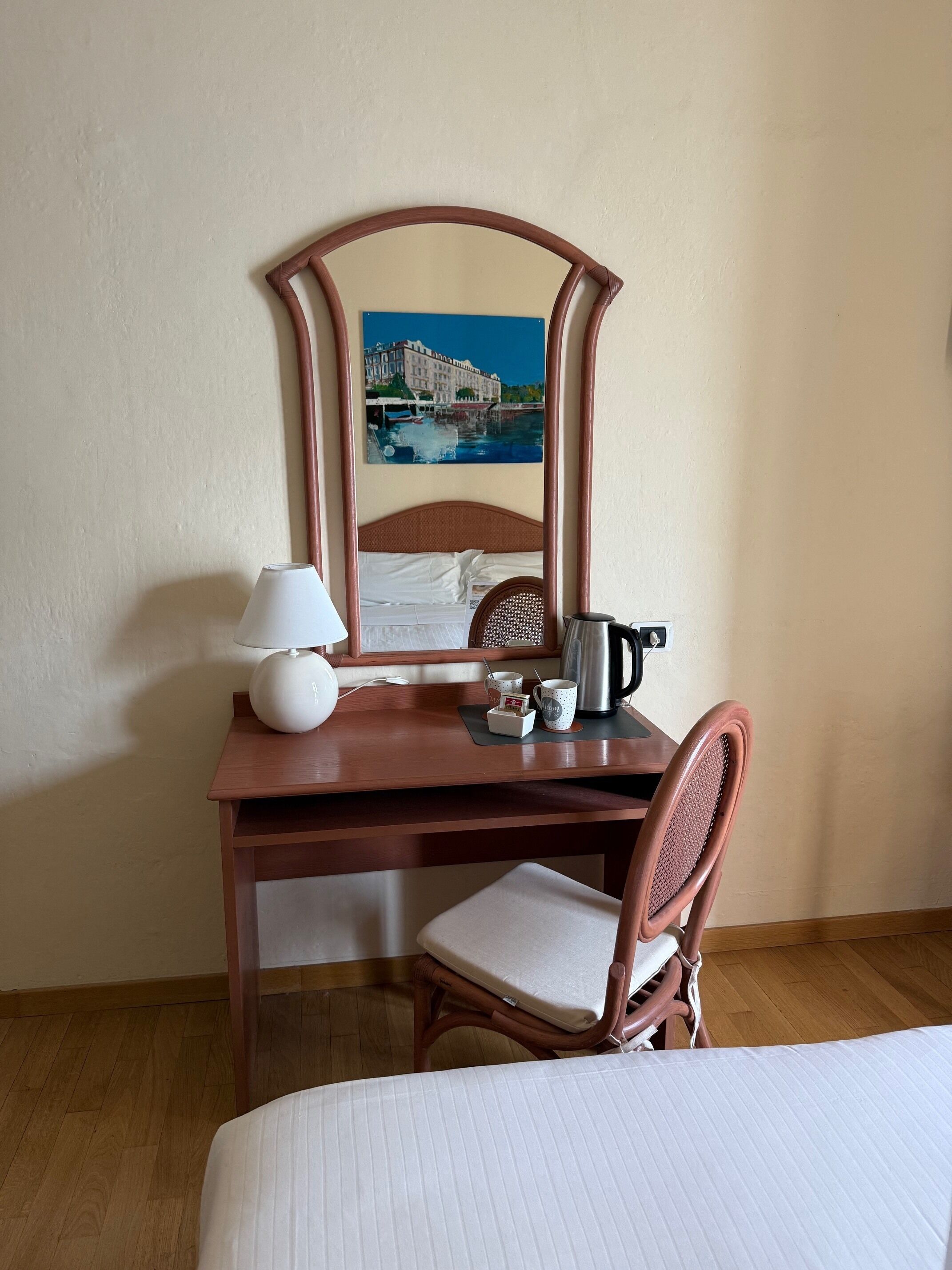 A hotel room with a wooden desk, a chair, a bed, a mirror, a painting, a lamp, a kettle, and a cup.