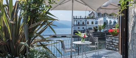 Terraza con mesa y sillas bajo un paraguas grande, con vistas a una masa de agua y un edificio con balcones.