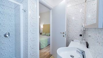 Une salle de bain avec une douche, un lavabo et un miroir. Une porte donne accès à une chambre.