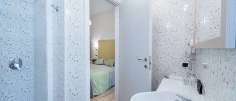A bathroom with a shower, a sink, and a mirror. There is a door leading to a bedroom.