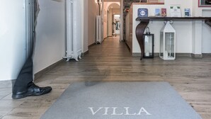 Zona de recepción de hotel con un felpudo que dice "VILLA BELVEDERE" y suelo de madera.