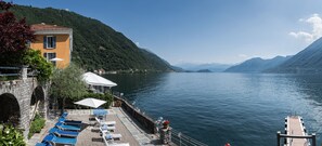 A lakeside hotel with a terrace, lounge chairs, and a stone wall.