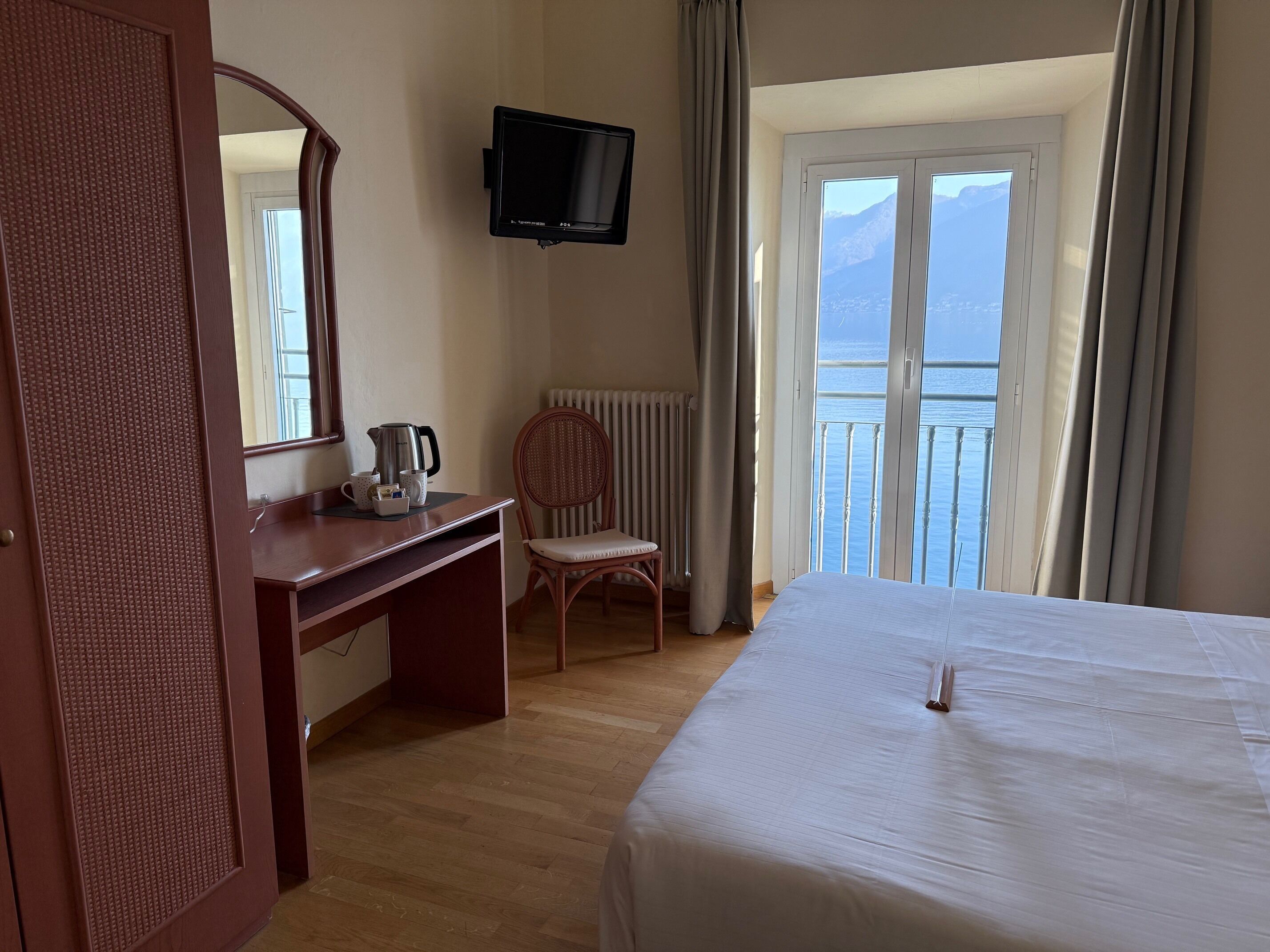 A hotel room with a bed, a desk with a kettle, a chair, a television, and a view of the sea.