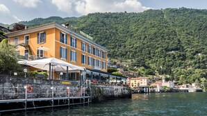Un edificio frente al agua con terraza de restaurante, un muro de piedra y vistas a una ladera verde.