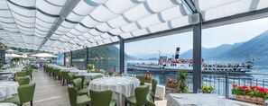 A restaurant with outdoor seating overlooking a lake, a boat with passengers, and mountains in the background.