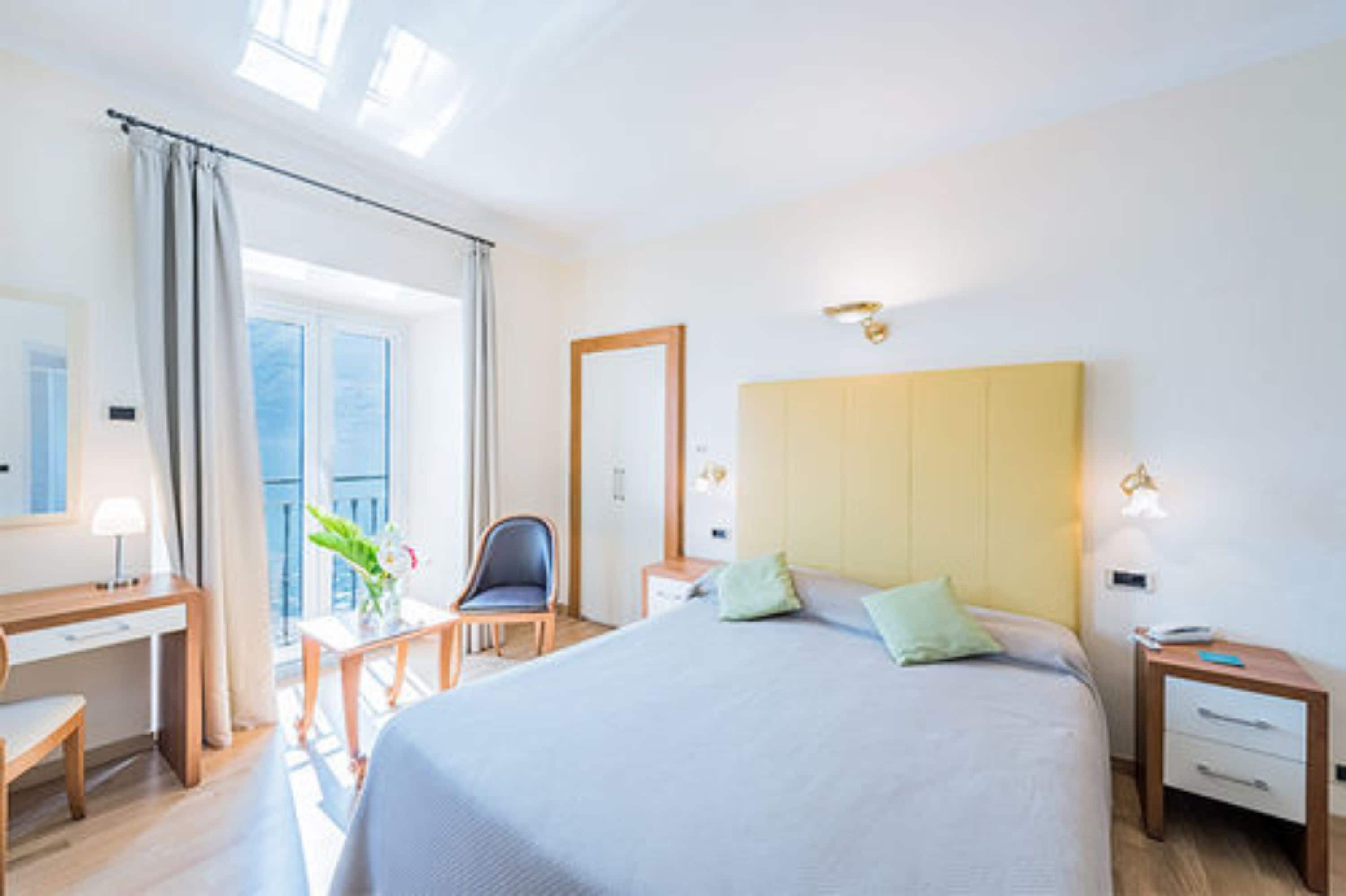 A hotel room with a large bed, a desk, a chair, a nightstand, and a window with curtains.