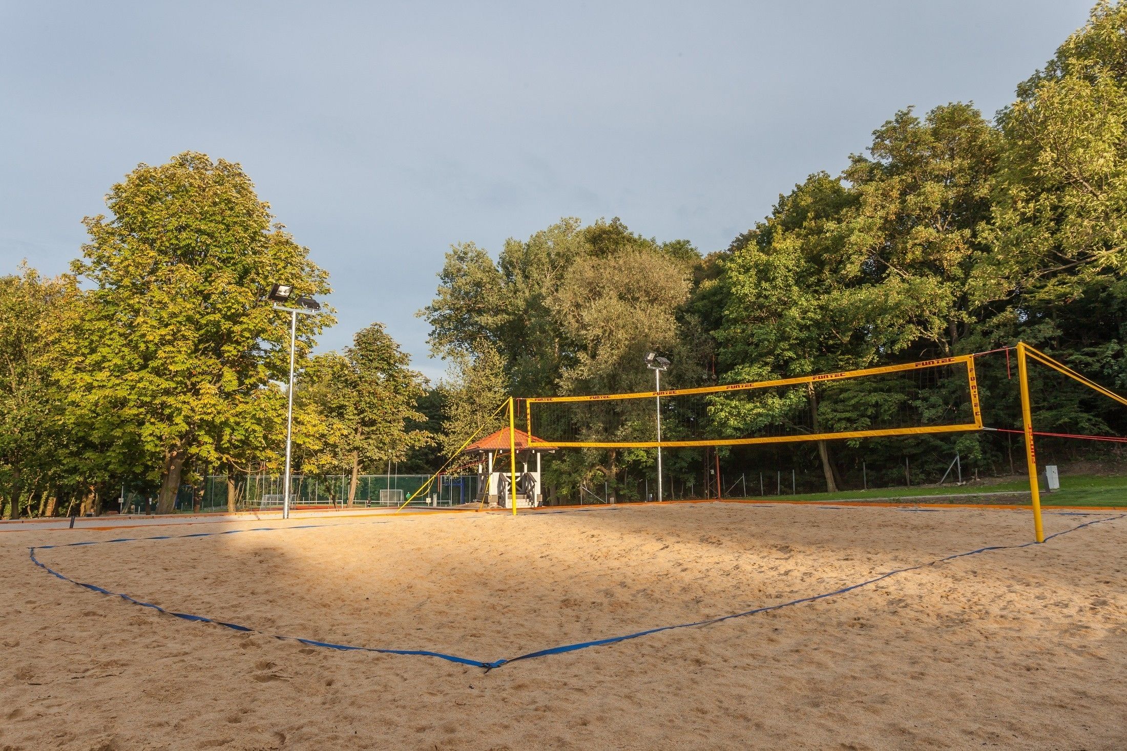 A volleyball court with a net, surrounded by trees and a small pavilion.