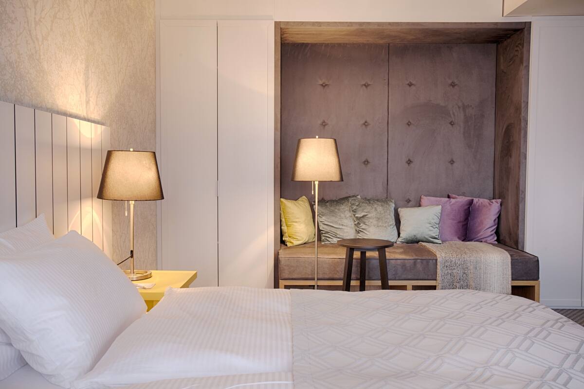 A hotel room with a bed, two bedside lamps, a sofa with cushions, and a small table.