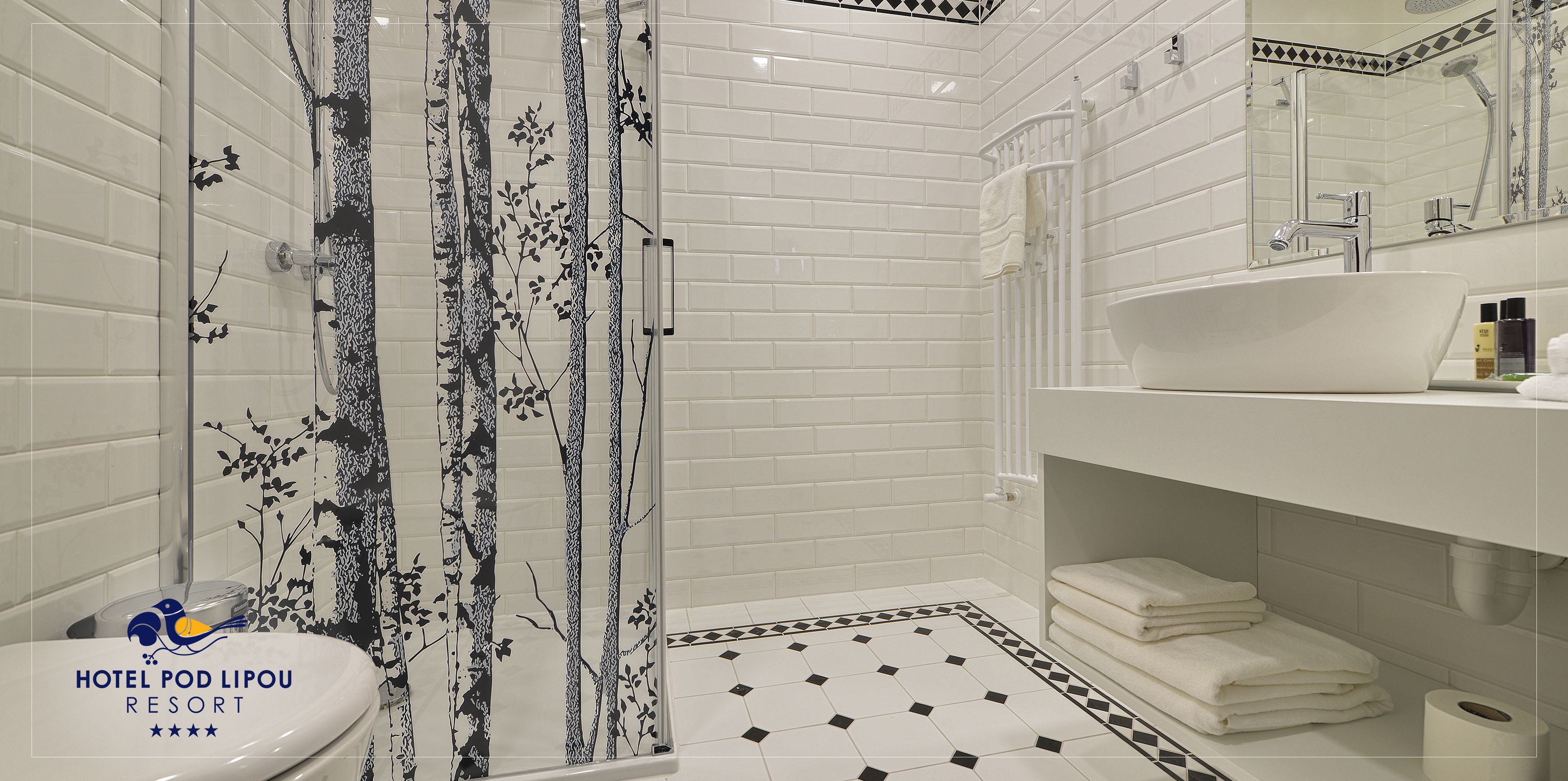 Uma casa de banho com azulejos brancos tipo metrô, um decalque de árvore em preto e branco, uma pia branca e zona de duche.