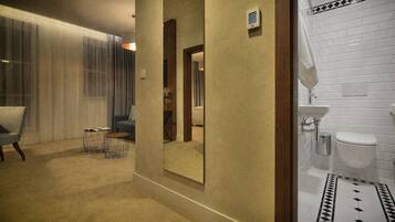 A hotel room with a dining area, a large mirror, a modern bathroom with white tiles, and a patterned rug.