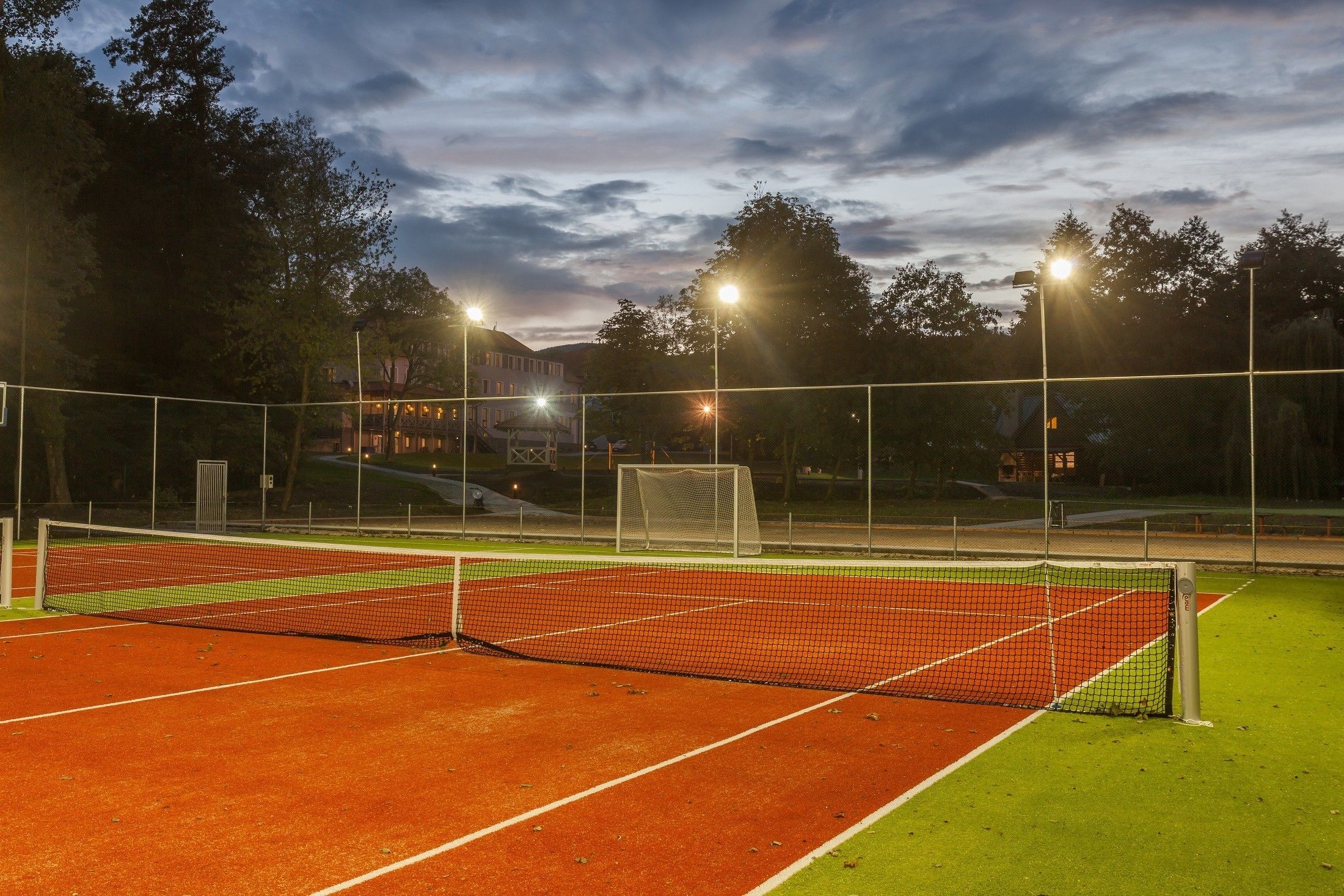 A tennis court with a net, surrounded by a fence, and illuminated by overhead lights.