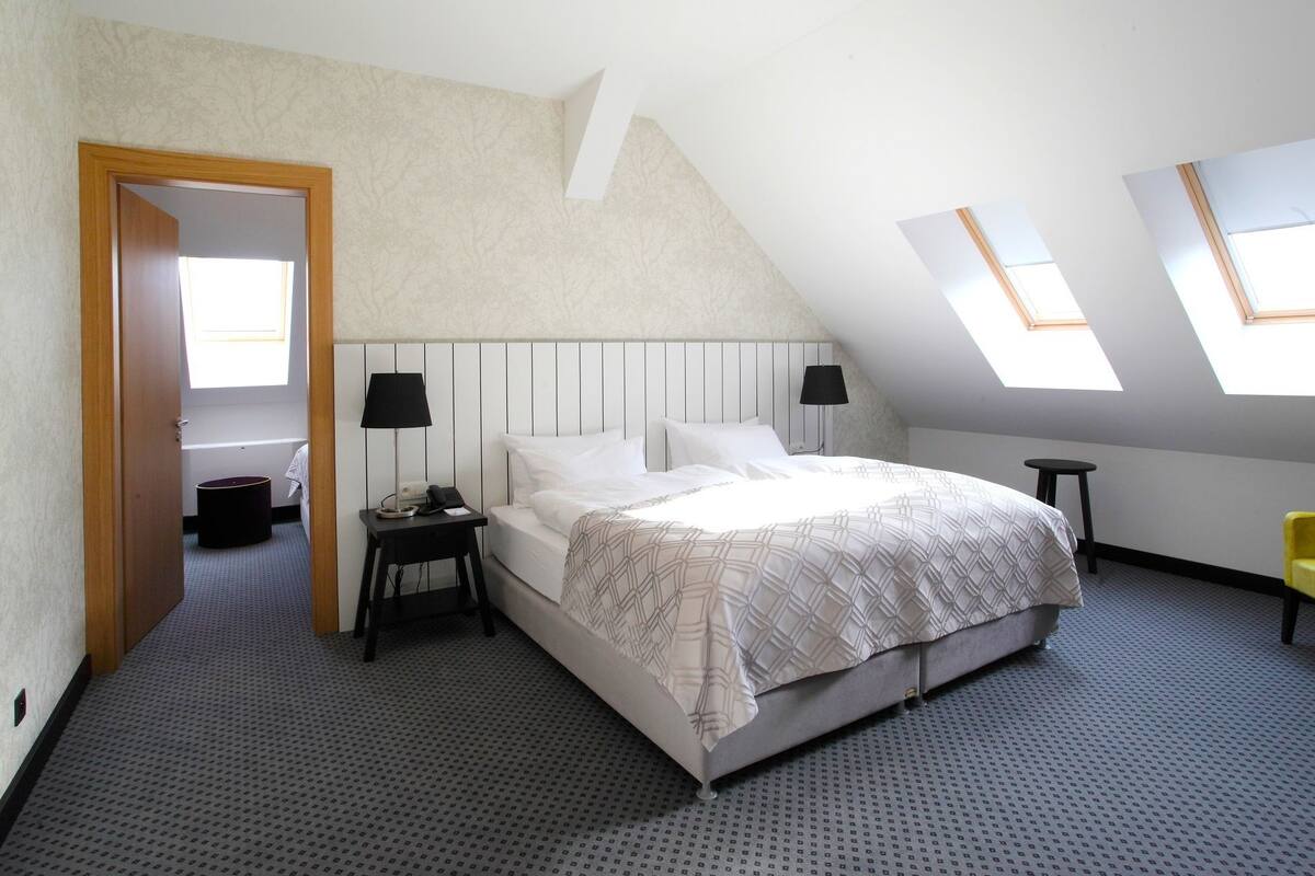 A bedroom with a bed, two bedside tables, a lamp, a small stool, and a door.
