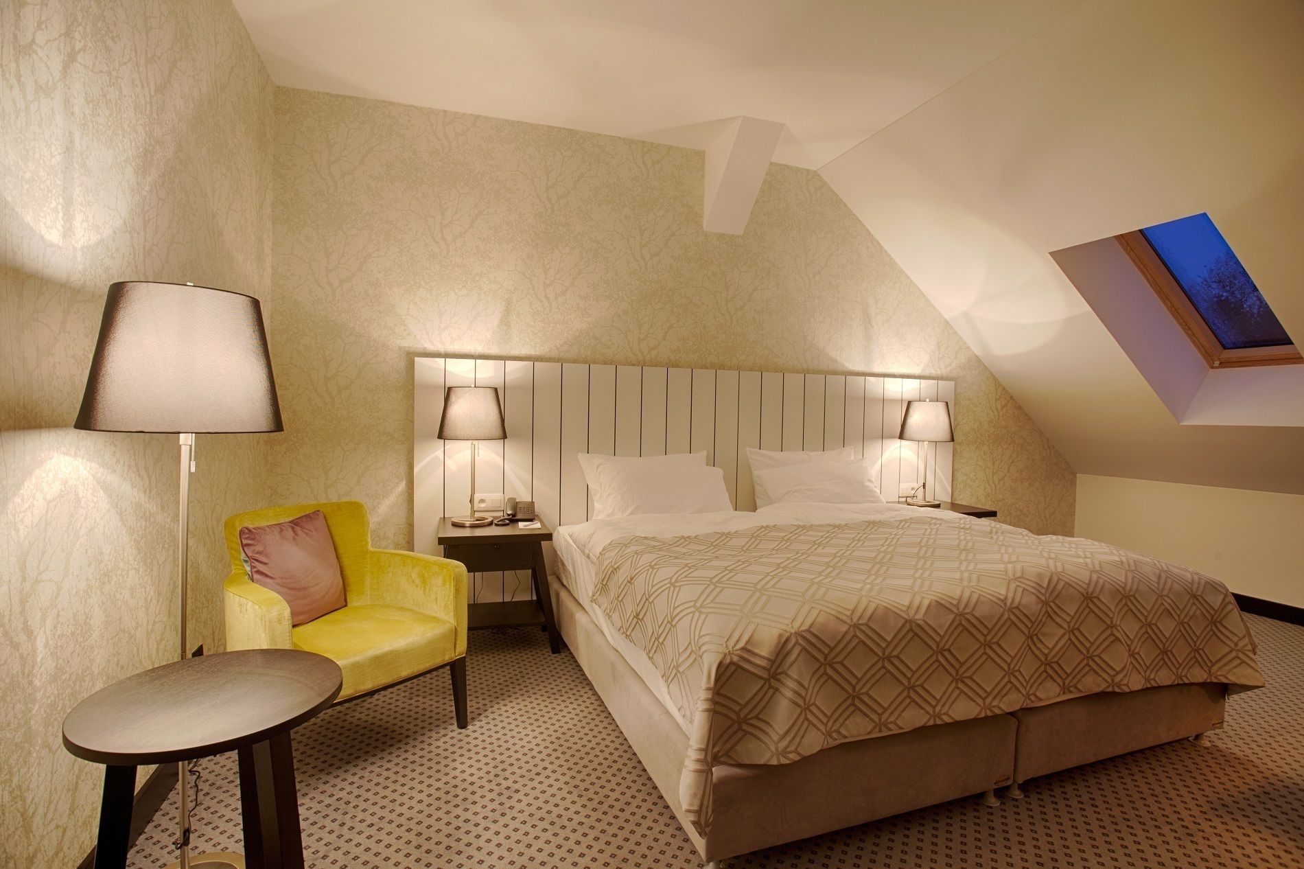 A hotel room with a bed, two bedside lamps, a small table, a yellow armchair, and a painting on the wall.