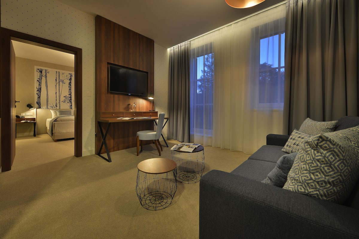 A modern hotel room with a sofa, two wireframe side tables, a desk with a chair, a flat-screen TV mounted on the wall, and a large window with curtains.