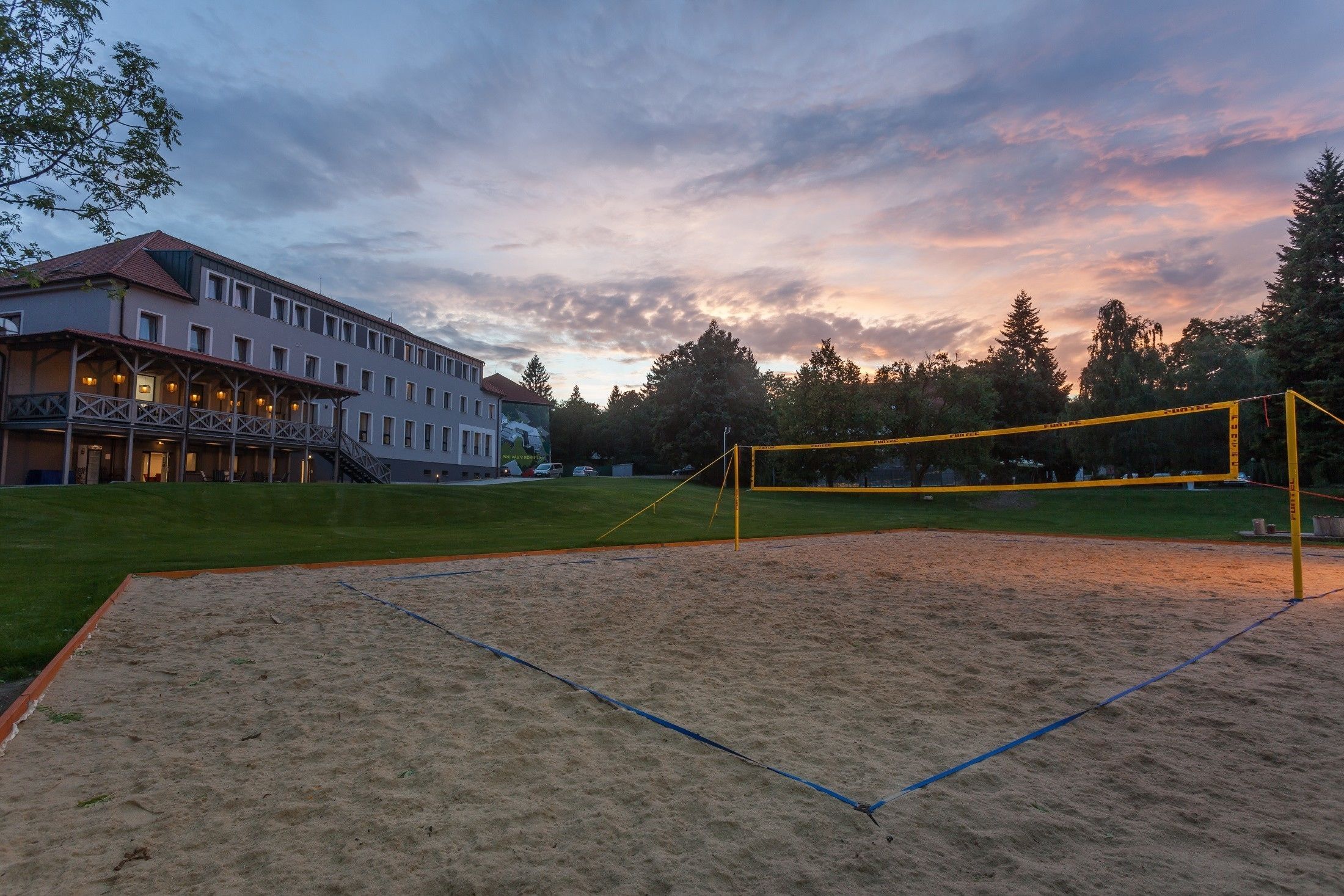 A sandy volleyball court with a net, surrounded by a grassy area and a building with multiple windows and balconies.