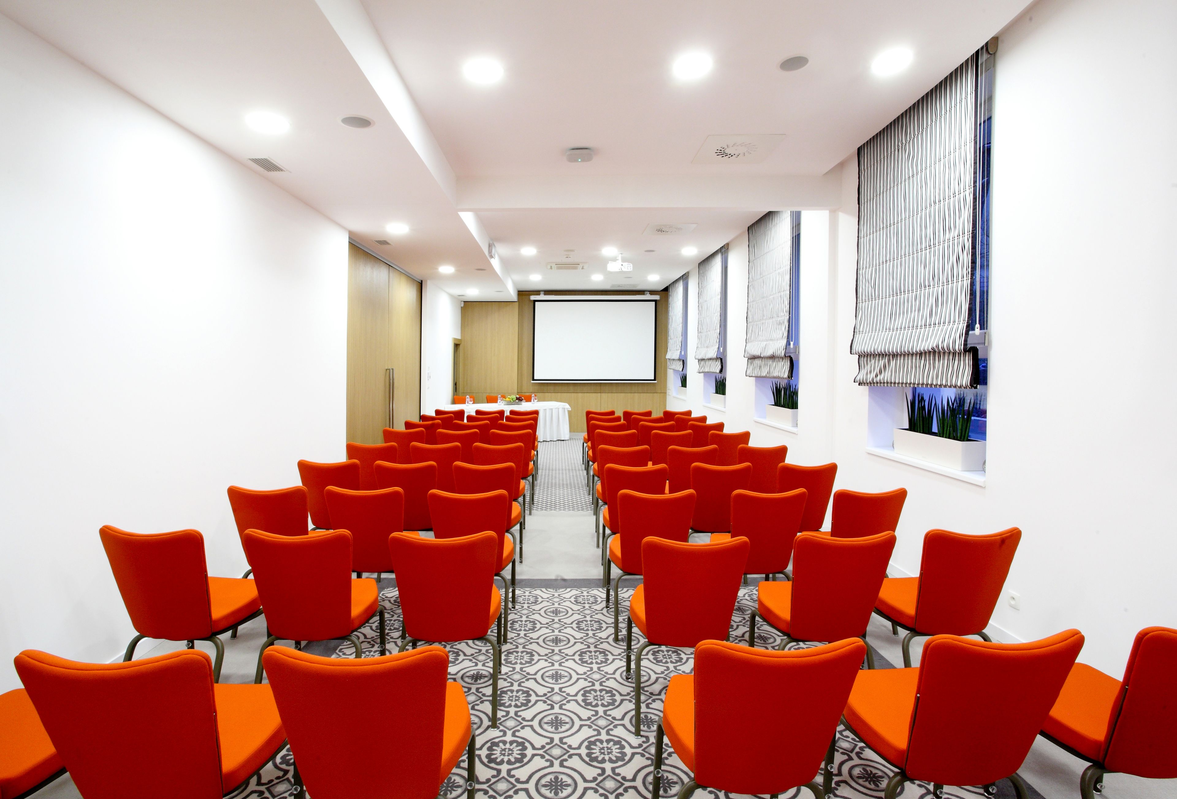 A conference room with rows of orange chairs, a projector screen, and patterned flooring.