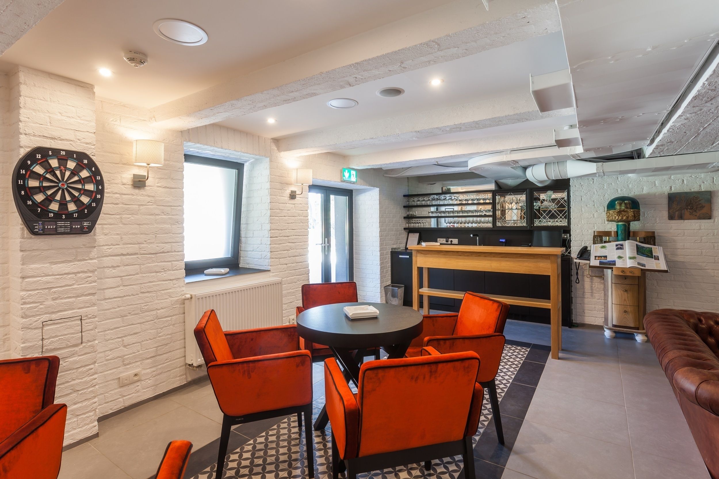 A modern lounge area with a dartboard, a round table with orange chairs, a fireplace, and a bar area.