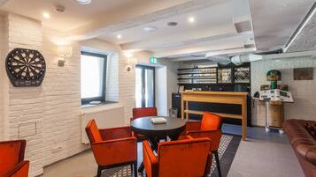 A modern lounge area with a dartboard, a round table with orange chairs, a fireplace, and a bar area.