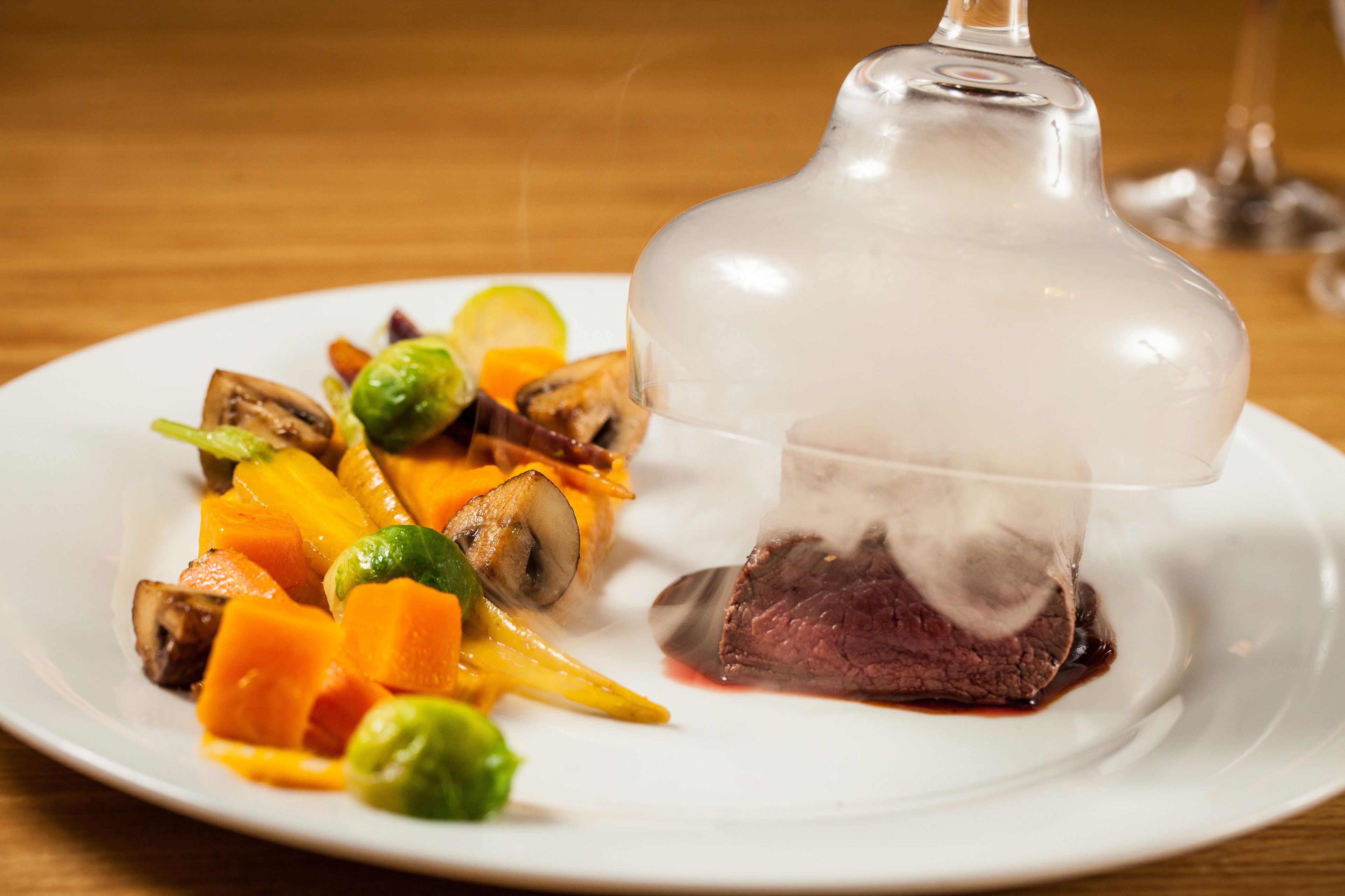 A white plate with a glass dome containing a piece of meat, surrounded by vegetables.