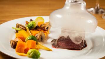 A white plate with a glass dome containing a piece of meat, surrounded by vegetables.