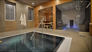 A modern indoor swimming pool with clear glass walls, wooden paneling, and a seating area with a view of the pool. - Hotel Pod Lipou (Modra)