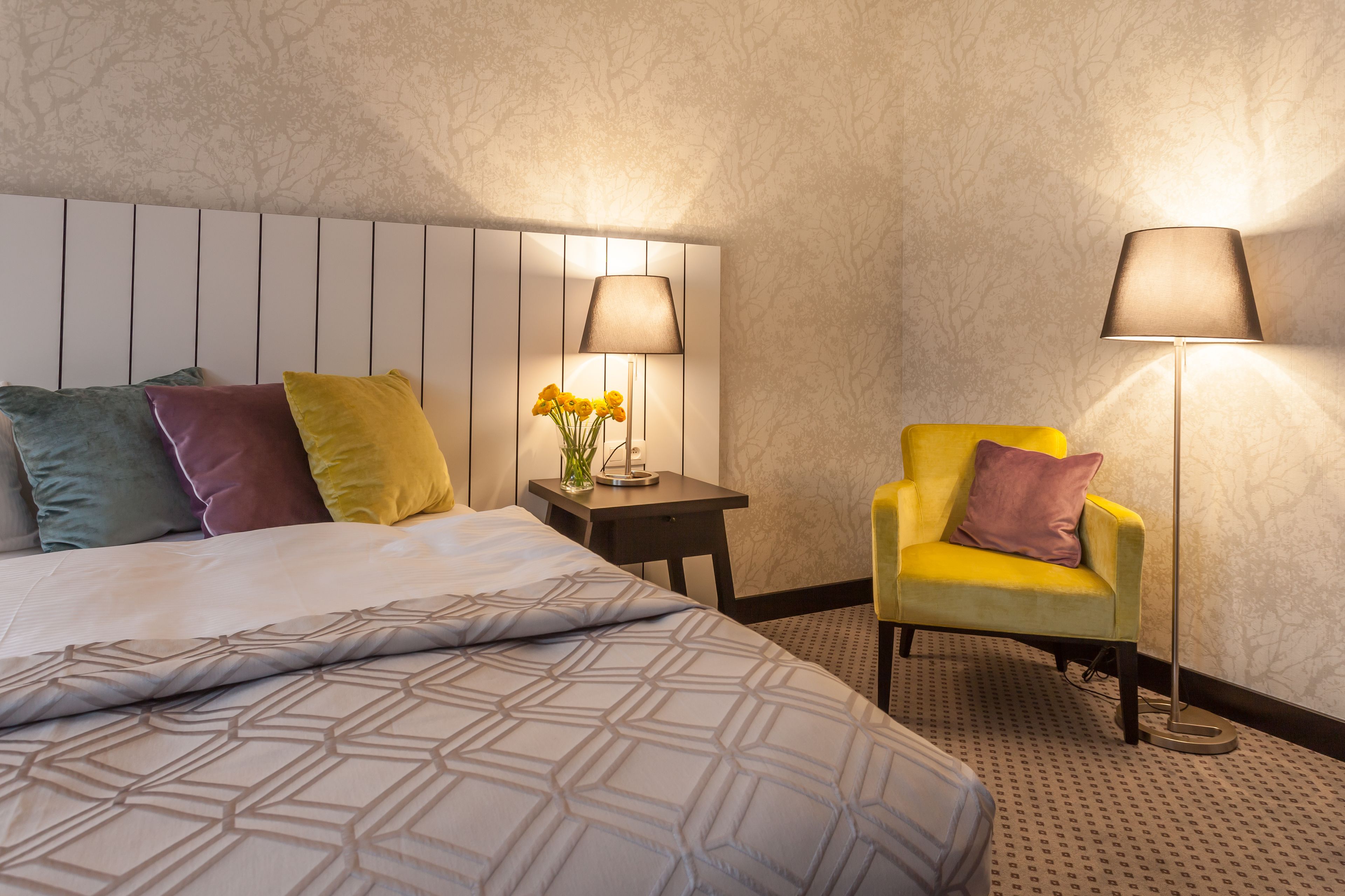 Habitación de hotel con una cama, una mesita de noche con lámpara, una silla y una pequeña mesa con flores.