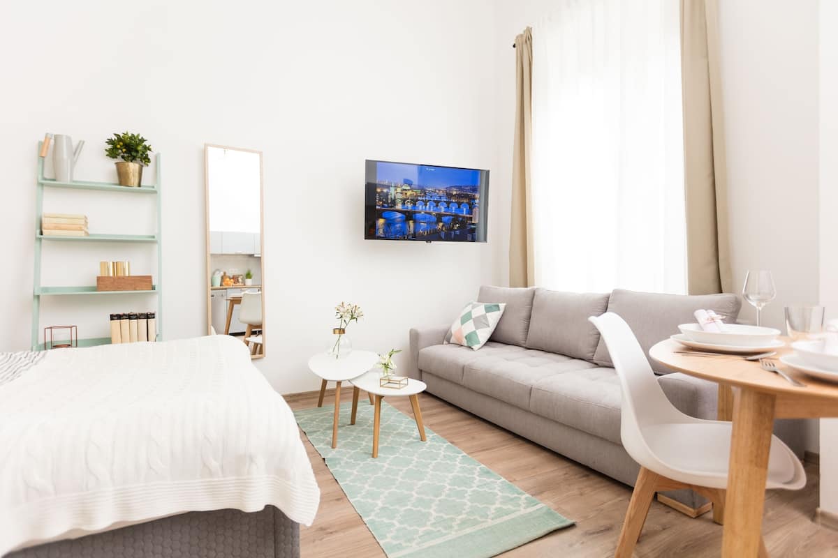 A modern hotel room with a bed, a sofa, a dining table, a TV, and a cityscape view.