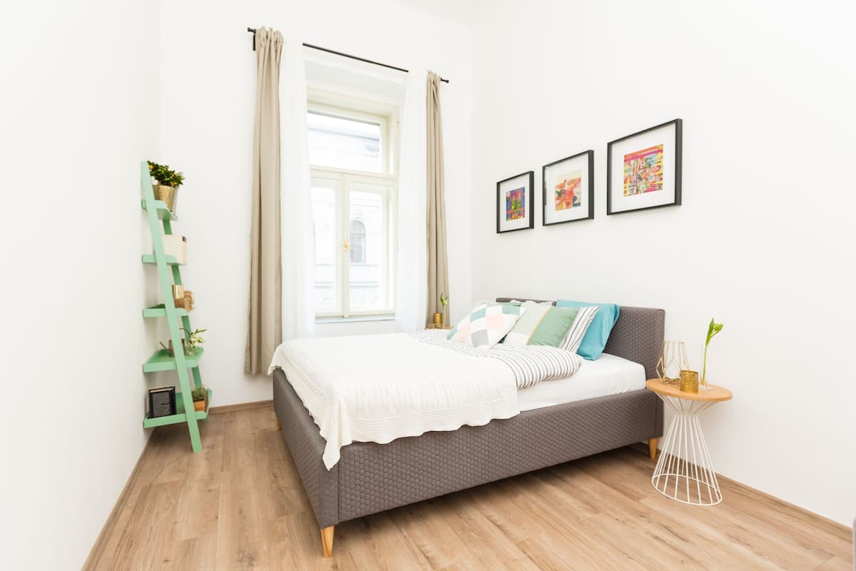 A bedroom with a bed, a window with curtains, a green ladder, and three framed pictures on the wall.