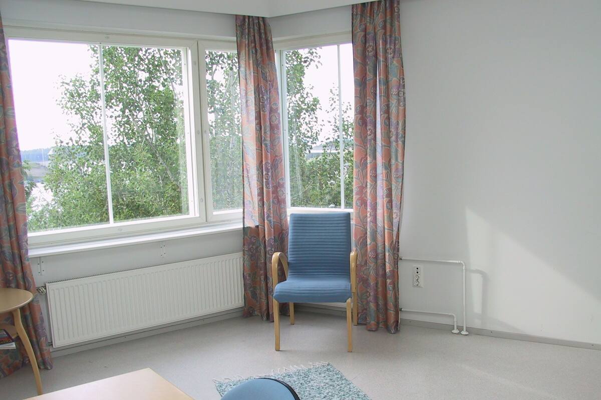 Huoneessa on suuri ikkuna, sininen tuoli, puinen pöytä ja kuviollinen matto.