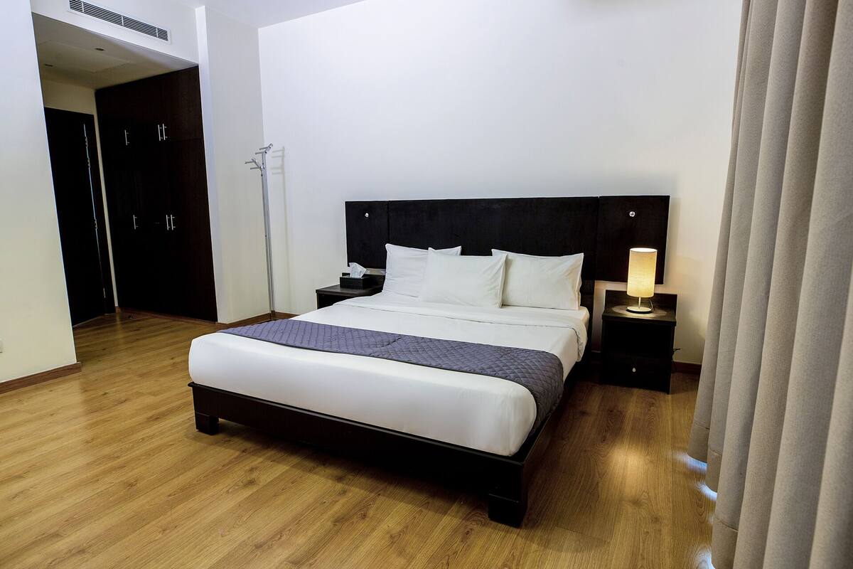 Une chambre d’hôtel avec un grand lit, une table de chevet avec une lampe, une armoire et un lampadaire.