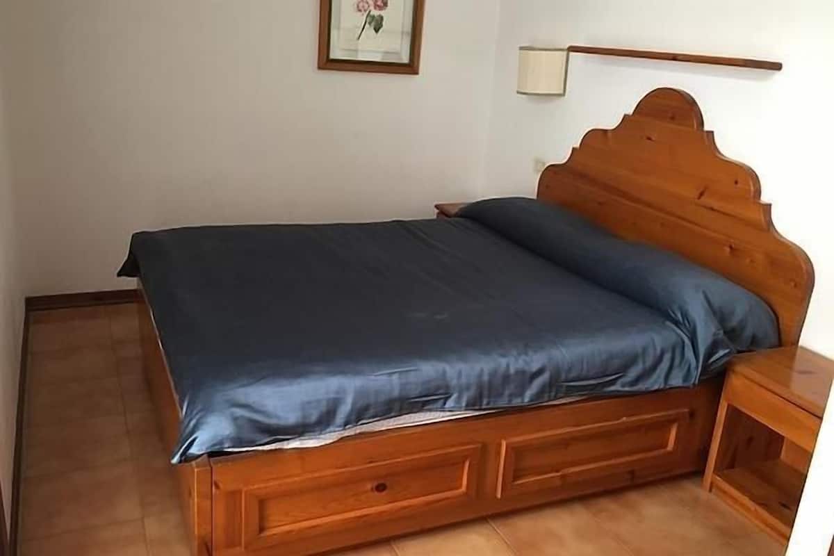 Una cama de madera con colchón y almohada azul oscuro a juego, una mesita de noche de madera y un cuadro en la pared.