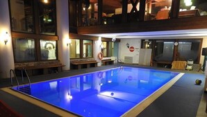 Ein großer, überdachter Innenpool mit blauer Färbung, umgeben von einer Holzterrasse und einer überdachten Sitzgruppe.