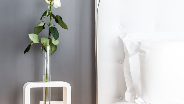 A modern bedroom with a white bed, a bedside table with a vase of a white rose, and a white bedside lamp. - M2M Made To Measure (Milan)