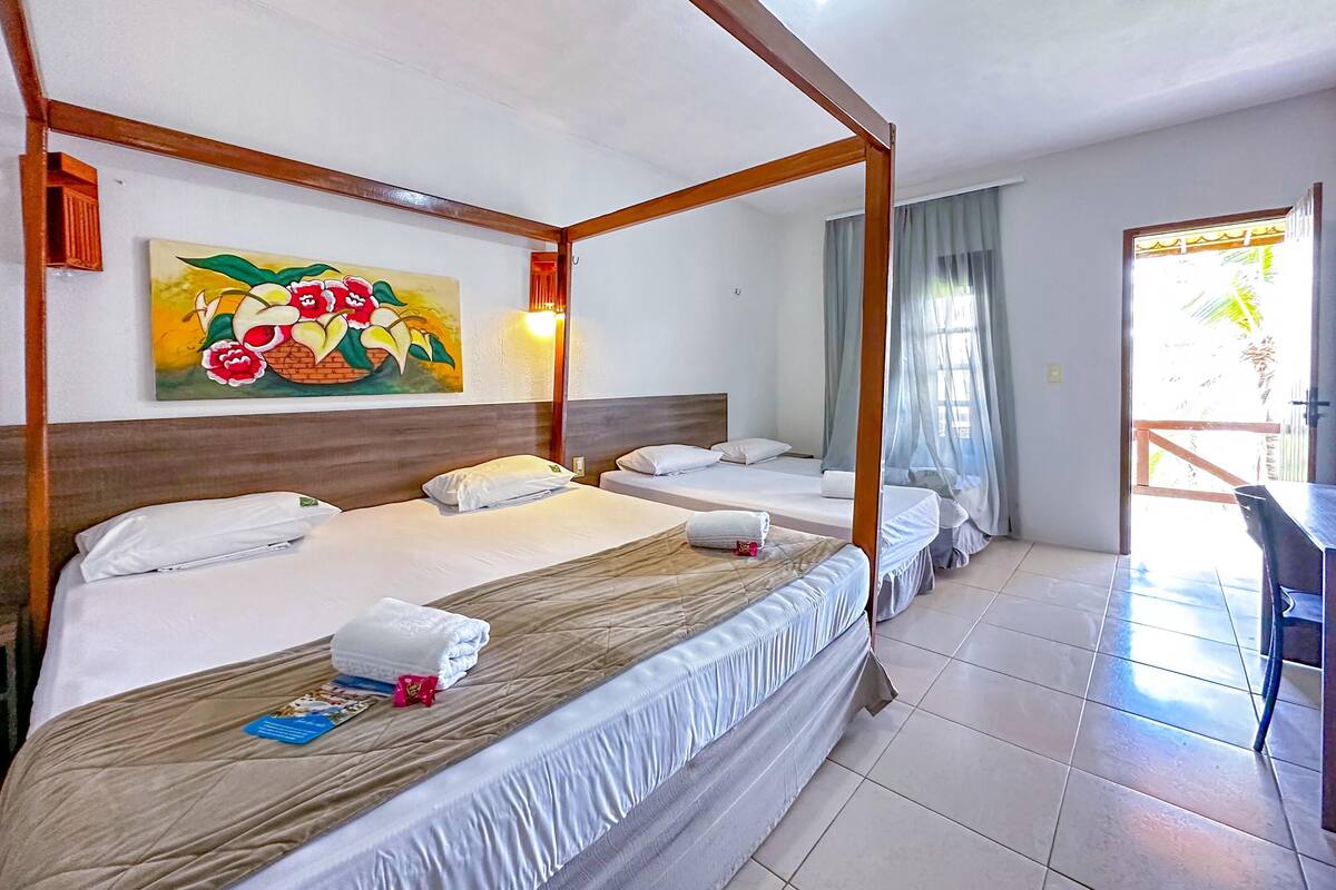 A hotel room with a four-poster bed, two single beds, a painting on the wall, and a view of the outdoors through a door.