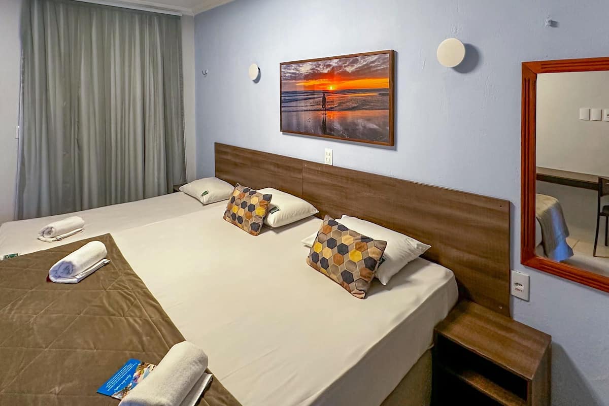A hotel room with a large bed, a painting of a sunset, a round wall clock, and a mirror.