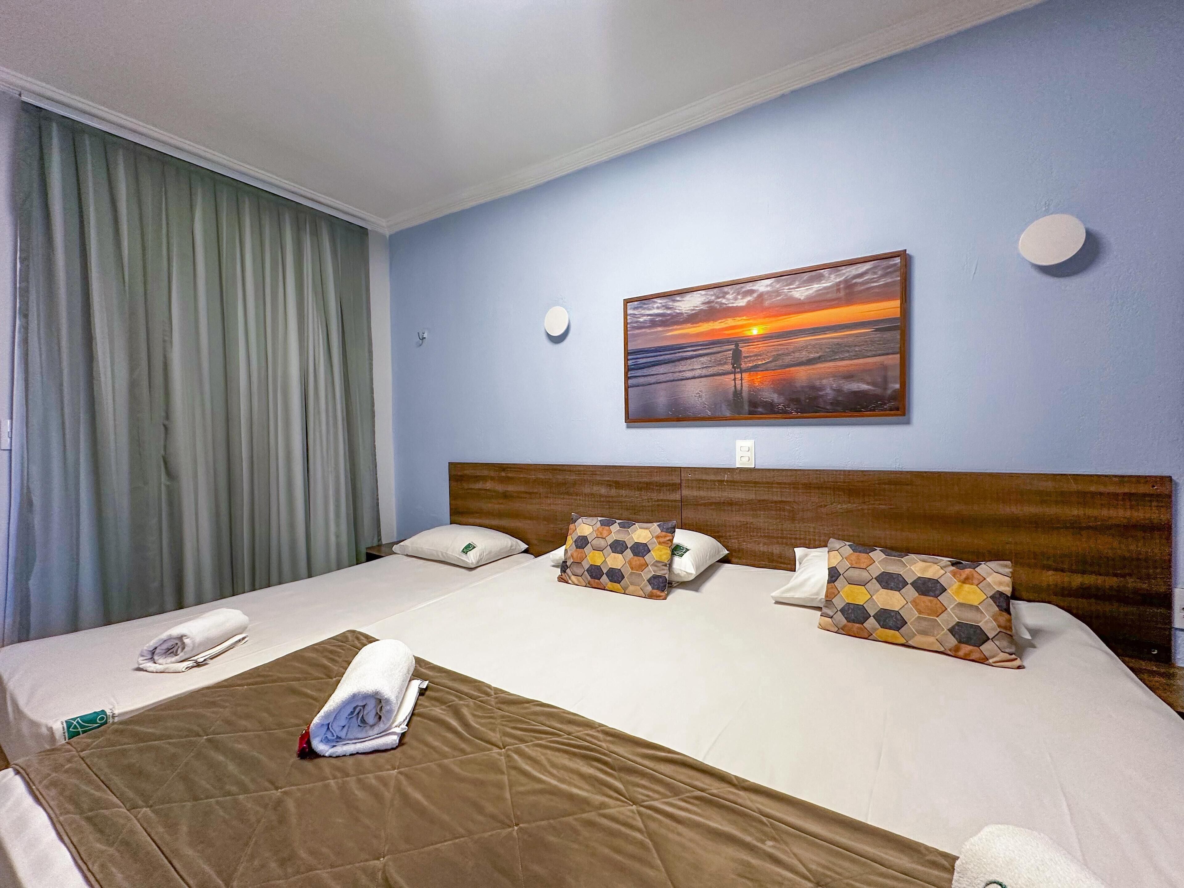 Une chambre d’hôtel avec un grand lit, deux oreillers, une couverture, une serviette, un tableau représentant un coucher de soleil, une fenêtre avec des rideaux et une table de chevet.