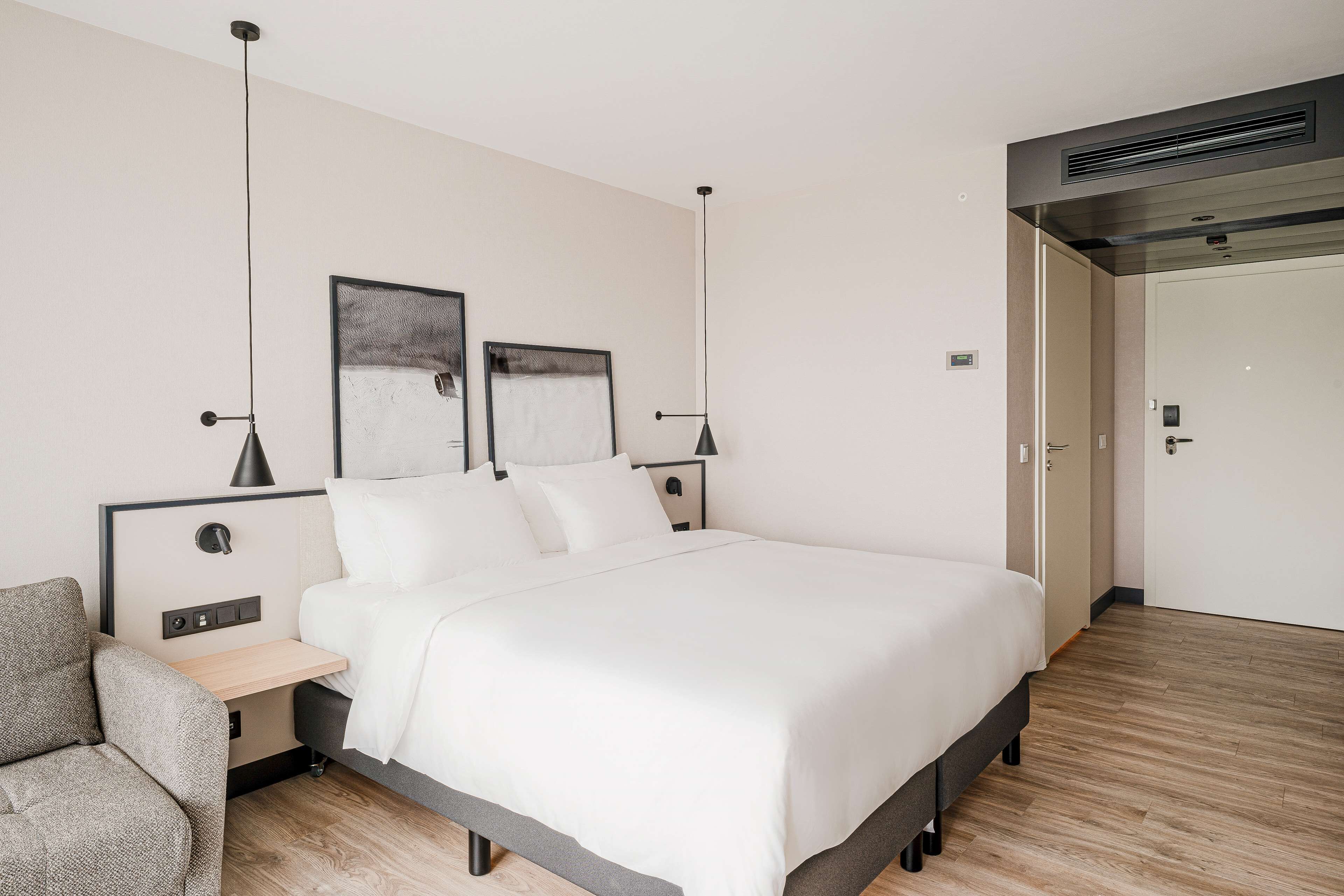 A modern hotel room with a large bed, bedside tables, a desk, and a chair. There are two wall-mounted lamps and a ceiling vent.