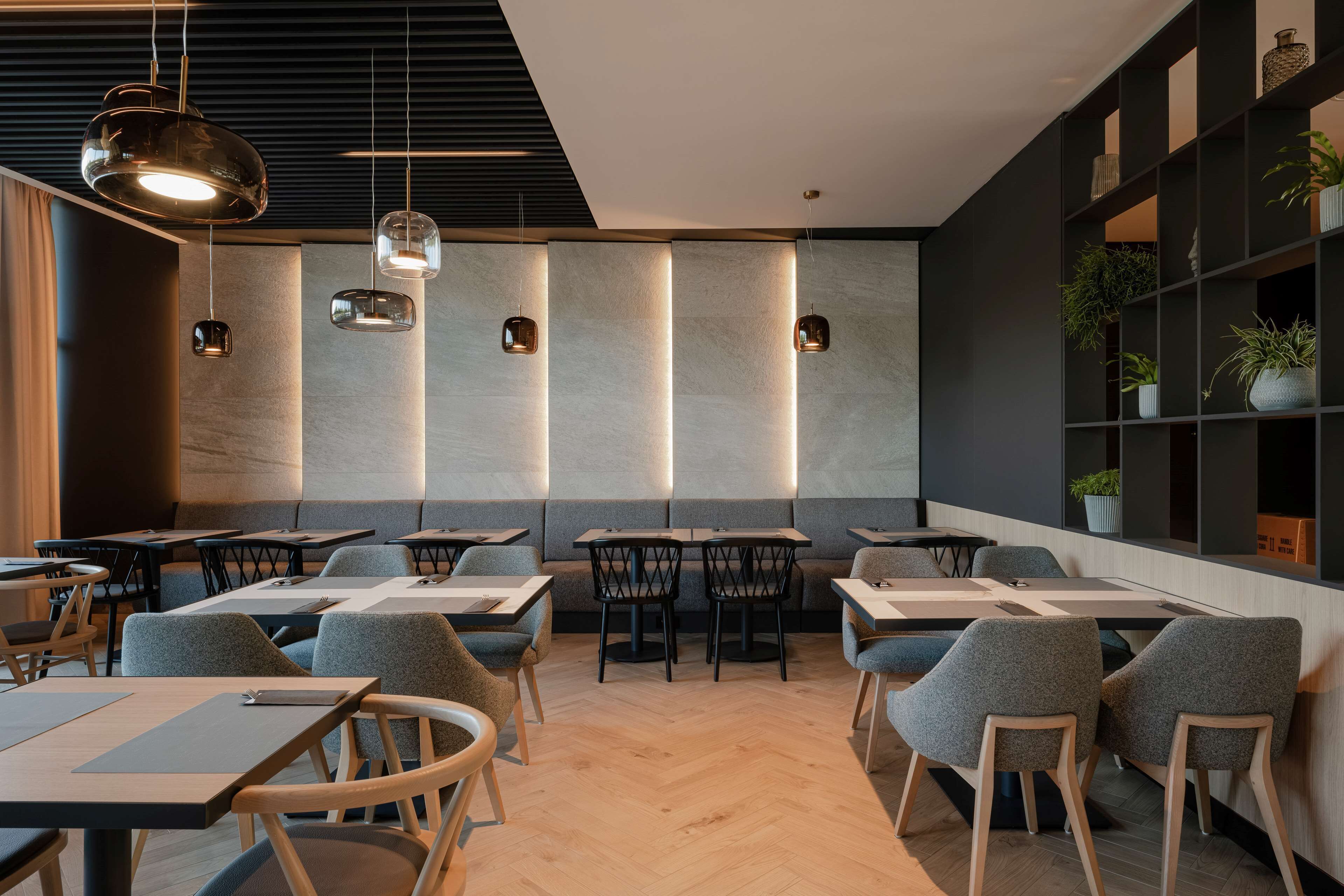 Et moderne restaurantmiljø med træborde, grå polstrede stole og hængende lamper.