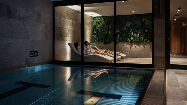 Een moderne binnenzwembadruimte met een glazen wand, een loungeruimte met uitzicht op een groene wand, en een bordje dat een sauna aangeeft.