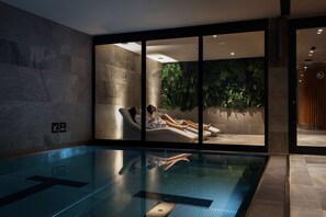 Een moderne binnenzwembadruimte met een glazen wand, een loungeruimte met uitzicht op een groene wand, en een bordje dat een sauna aangeeft.