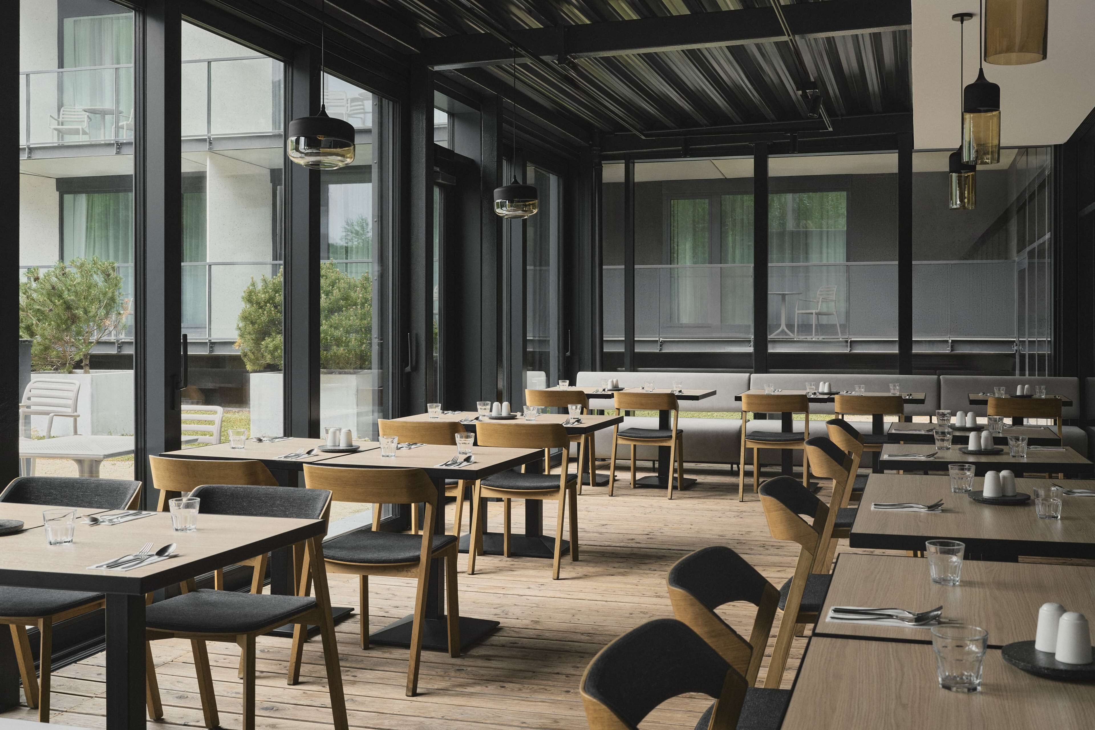 Un restaurant moderne avec des tables et des chaises en bois, de grandes fenêtres et des lampes suspendues.