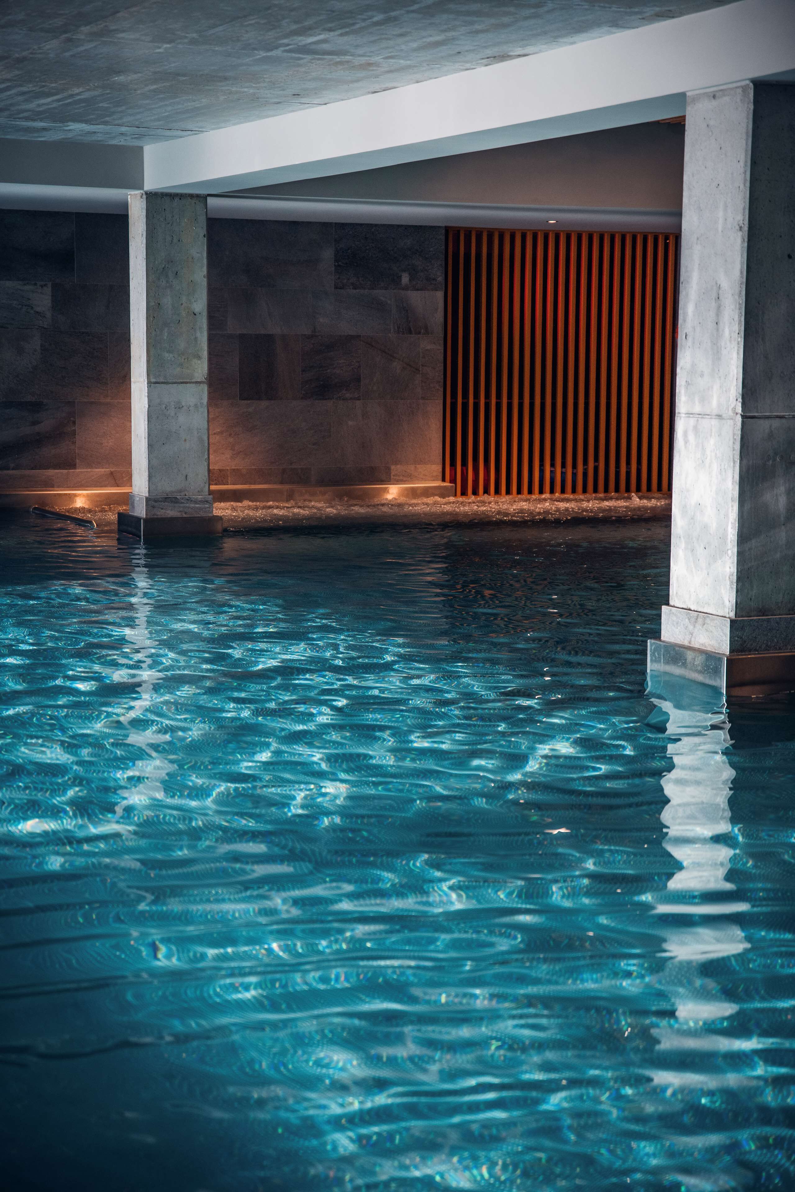 An indoor swimming pool with clear water, concrete pillars, and a wooden slatted wall in the background.