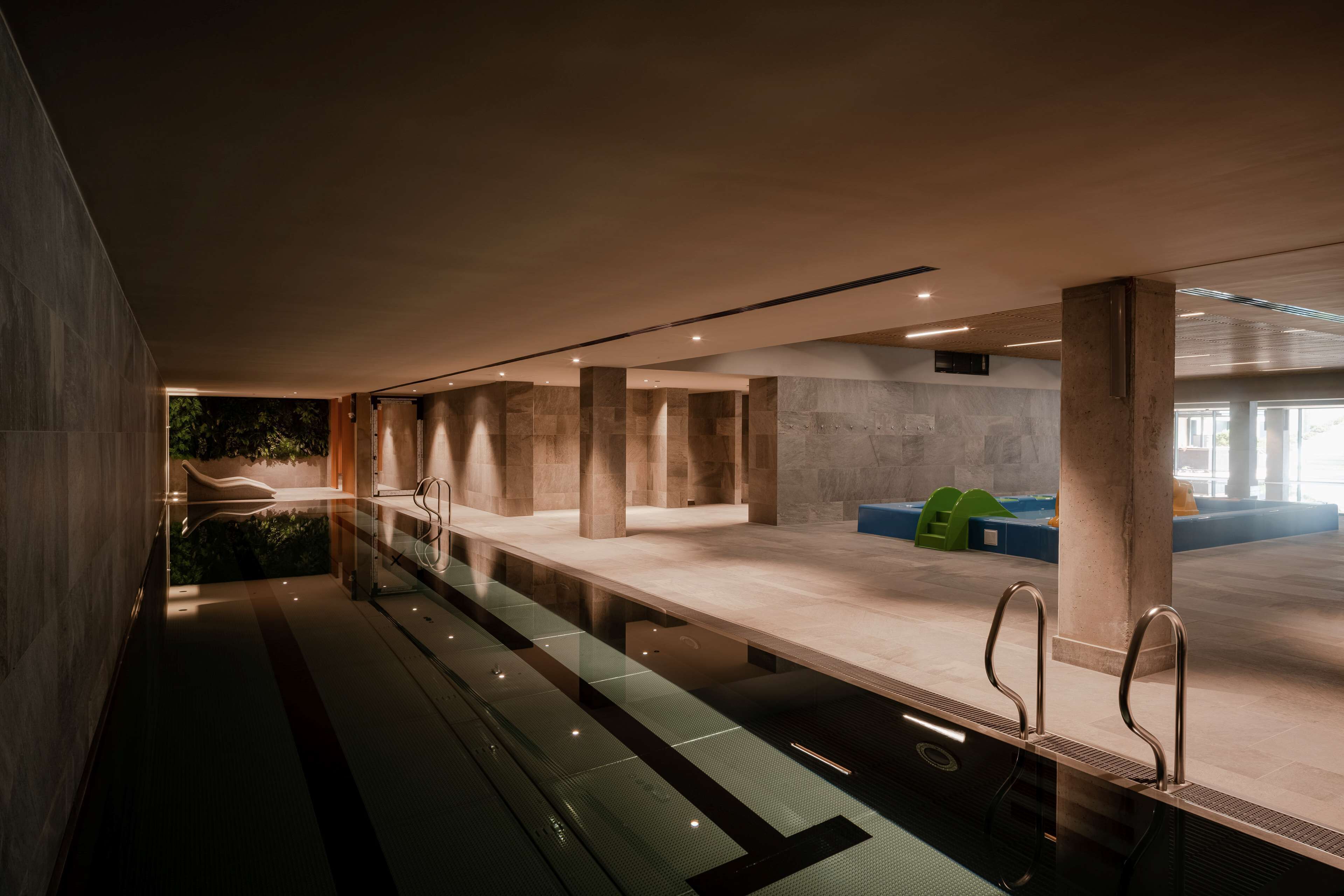 A modern indoor swimming pool with a glass barrier, concrete pillars, and a view of greenery outside.