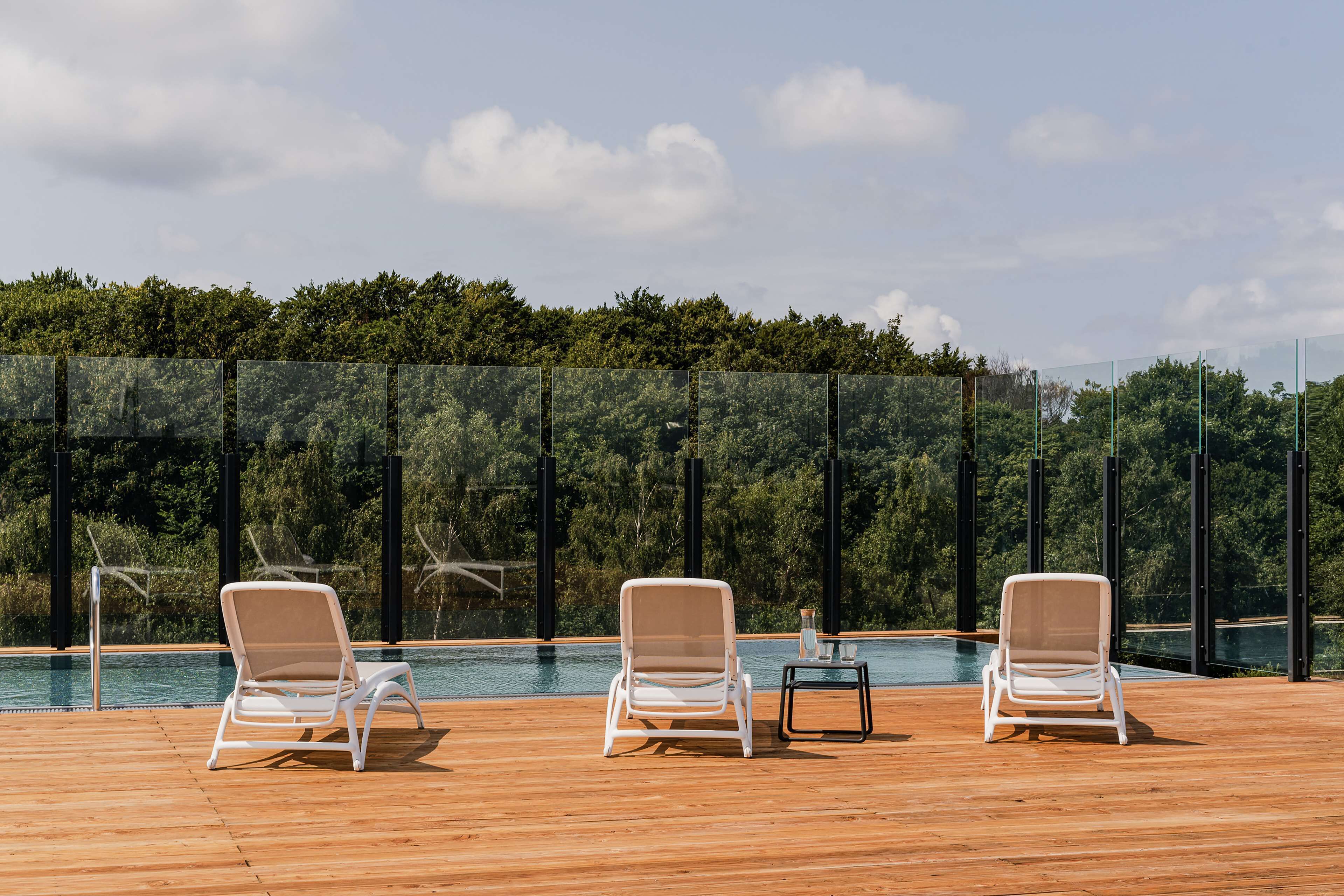 A poolside area with three white lounge chairs and a small table, surrounded by a wooden deck and a glass fence.