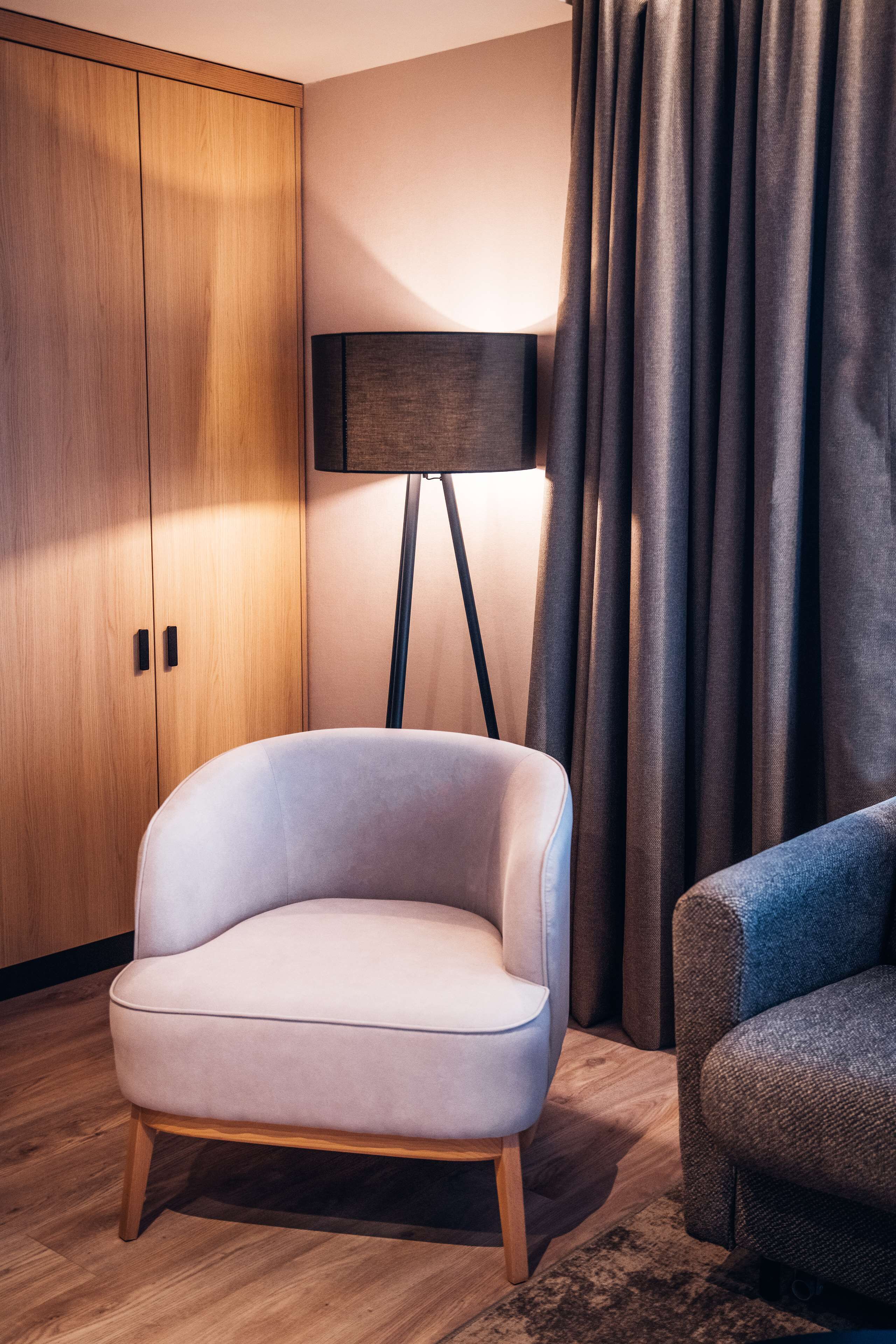 Ein modernes Zimmer mit einem hellen Ohrensessel, einer dunklen Stehlampe und einem Holzschrank.