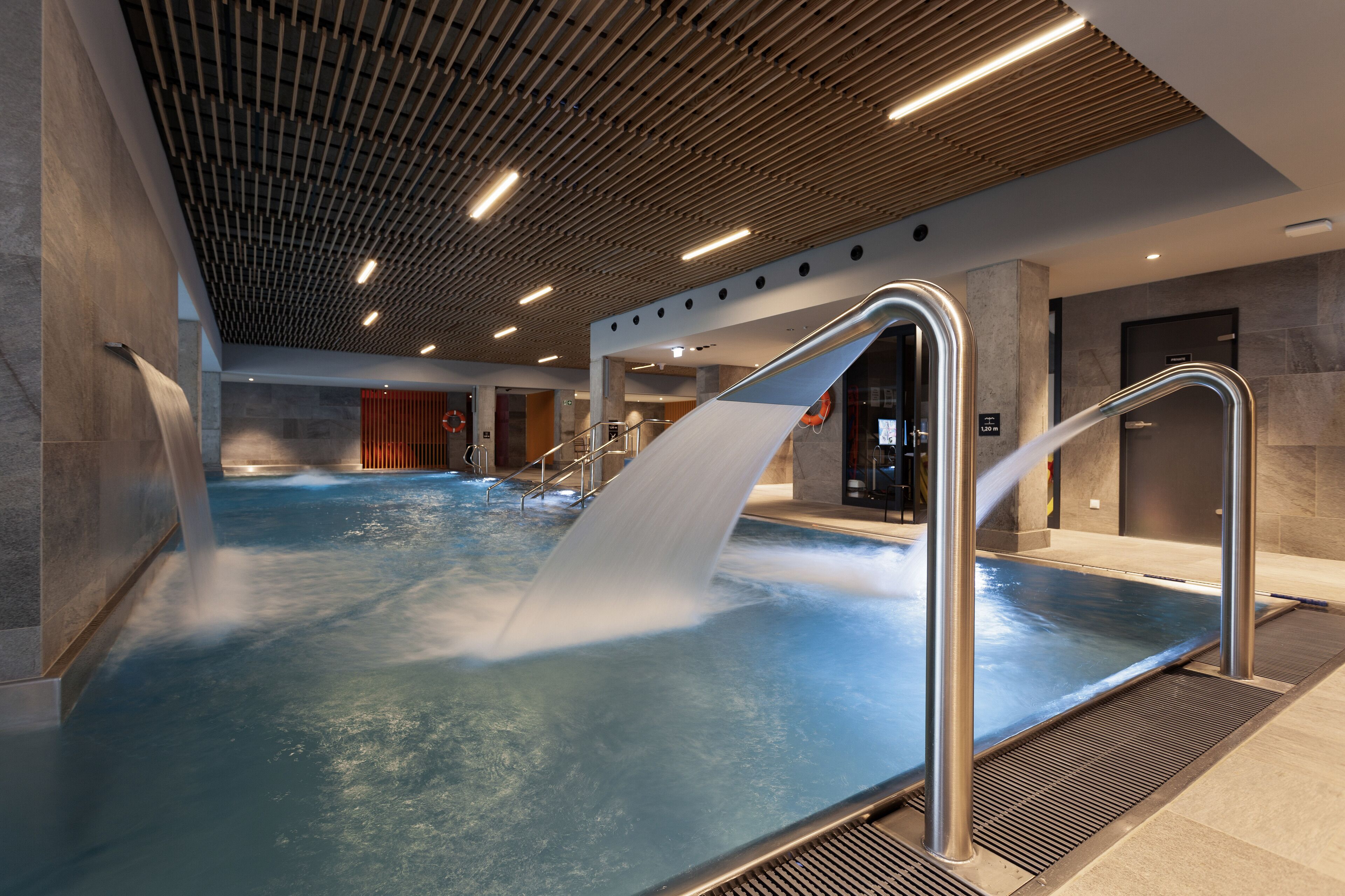 A modern indoor pool with a wooden ceiling, metal railings, and a visible exit door.