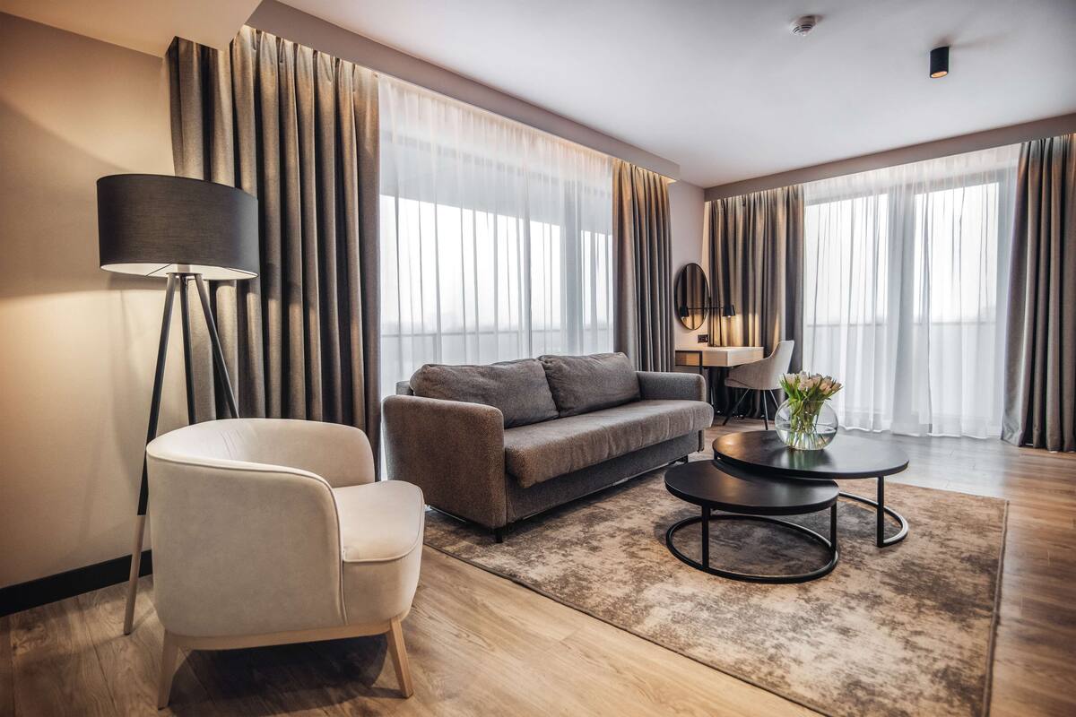 A modern living room with a sofa, armchair, and coffee table, large windows with curtains, and a floor lamp.
