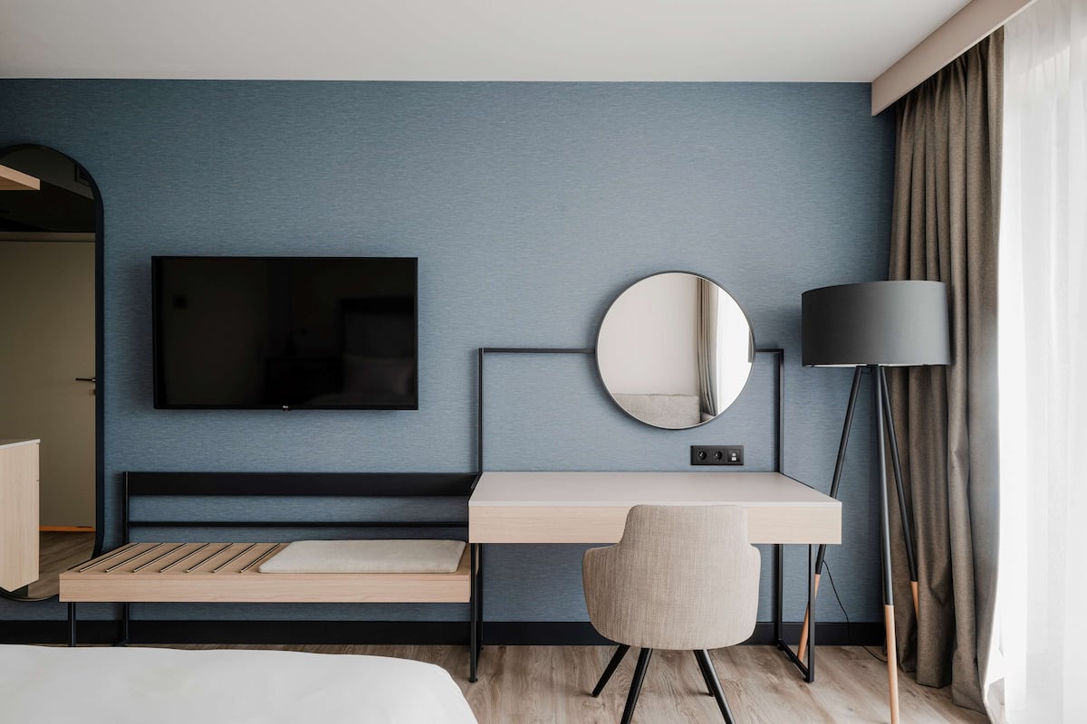 A modern hotel room with a bed, a desk, a chair, a TV, a round mirror, and a lamp.